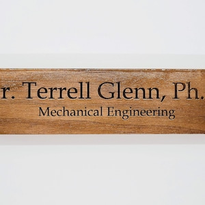 May include: A rectangular wooden desk sign with the engraved text "Dr. Terrell Glenn, Ph.D Mechanical Engineering." The sign is brown with silver-colored circular accents on each end.