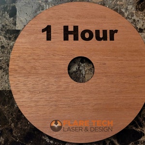 Peut inclure: Un disque rond en bois avec un trou au centre. Le disque est brun clair et porte le texte "1 Hour" grav&eacute; en lettres noires. Le bas du disque porte le texte "FLARE TECH LASER & DESIGN" en lettres noires.