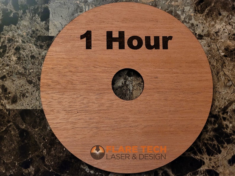 Peut inclure: Un disque rond en bois avec un trou au centre. Le disque est brun clair et porte le texte "1 Hour" grav&eacute; en lettres noires. Le bas du disque porte le texte "FLARE TECH LASER & DESIGN" en lettres noires.