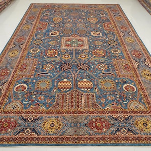 May include: A large, rectangular rug with an intricate floral design. The rug features a blue background with red, yellow, and cream-colored floral patterns. The border is a darker blue with repeating floral motifs. The rug is likely made of wool.