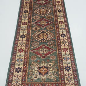 May include: A long, rectangular rug with a central teal field and intricate geometric patterns in red, beige, and navy blue. The rug has a border with floral motifs and measures approximately 3 feet wide.