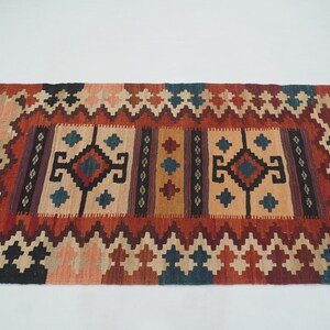 Puede incluir: Alfombra rectangular con un patr&oacute;n geom&eacute;trico en tonos rojos, beige, negro y azul. La alfombra presenta un dise&ntilde;o central con formas de diamantes y cuadrados, rodeado por un borde en zigzag.