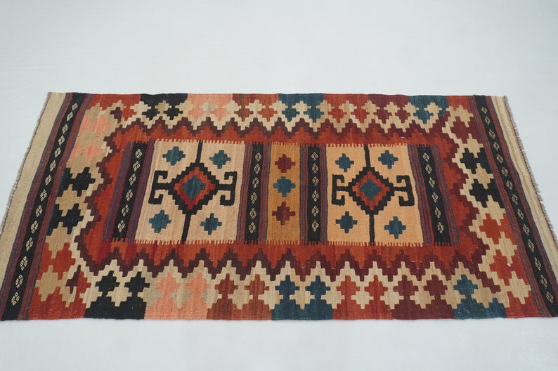 Puede incluir: Alfombra rectangular con un patr&oacute;n geom&eacute;trico en tonos rojos, beige, negro y azul. La alfombra presenta un dise&ntilde;o central con formas de diamantes y cuadrados, rodeado por un borde en zigzag.