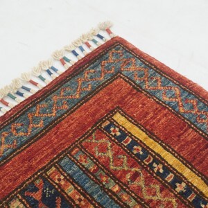 Może przedstawiać: Zbliżenie rogu czerwonego, niebieskiego i ż&oacute;łtego dywanu we wzory. Dywan ma geometryczne wzory i frędzelkową krawędź. Paleta kolor&oacute;w obejmuje odcienie czerwieni, błękitu, ż&oacute;łci i kremu.