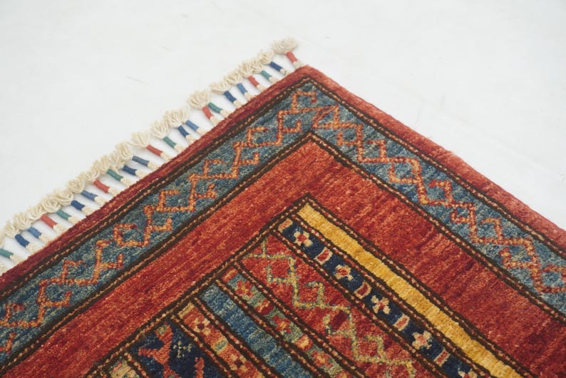 Może przedstawiać: Zbliżenie rogu czerwonego, niebieskiego i ż&oacute;łtego dywanu we wzory. Dywan ma geometryczne wzory i frędzelkową krawędź. Paleta kolor&oacute;w obejmuje odcienie czerwieni, błękitu, ż&oacute;łci i kremu.