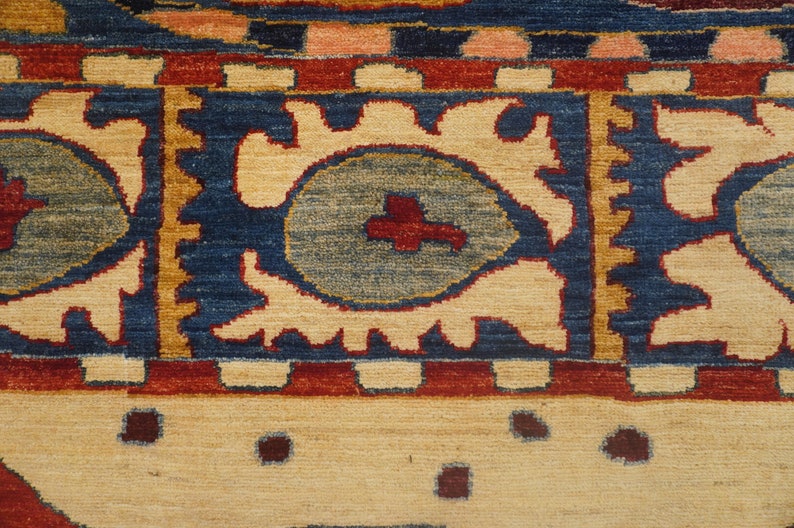 Op de afbeelding: Close-up van een tapijt met een blauwe achtergrond en een cr&egrave;mekleurig patroon. Het patroon bevat gestileerde bloemmotieven en een centraal medaillon met een rood kruis.