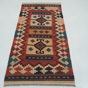 Puede incluir: Alfombra rectangular con un dise&ntilde;o central de formas geom&eacute;tricas en beige, rojo, negro y azul. La alfombra tiene un borde con un patr&oacute;n de diamantes repetido en varios colores. La alfombra est&aacute; hecha de material tejido.