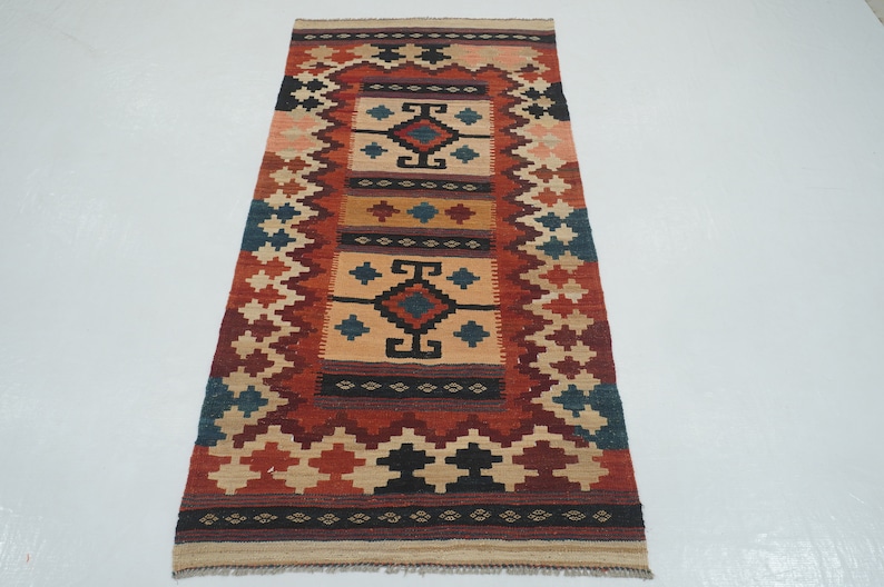 Puede incluir: Alfombra rectangular con un dise&ntilde;o central de formas geom&eacute;tricas en beige, rojo, negro y azul. La alfombra tiene un borde con un patr&oacute;n de diamantes repetido en varios colores. La alfombra est&aacute; hecha de material tejido.