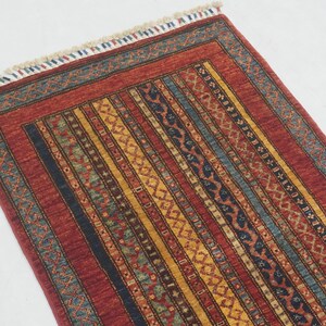 Może przedstawiać: Prostokątny dywan z czerwonym tłem i pionowymi paskami w kolorze ż&oacute;łtym, niebieskim i czarnym. Dywan ma wzorzystą obw&oacute;dkę i frędzle u g&oacute;ry. Wz&oacute;r jest skomplikowany i szczeg&oacute;łowy, odpowiedni do wystroju wnętrz.