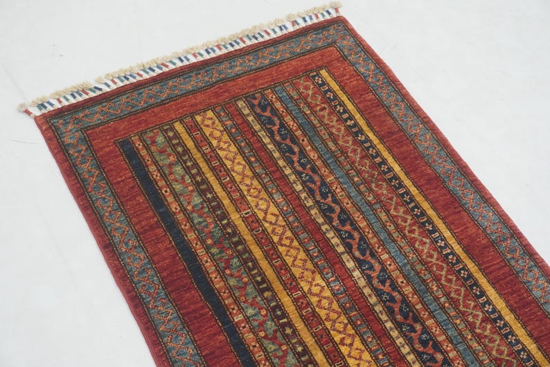 Może przedstawiać: Prostokątny dywan z czerwonym tłem i pionowymi paskami w kolorze ż&oacute;łtym, niebieskim i czarnym. Dywan ma wzorzystą obw&oacute;dkę i frędzle u g&oacute;ry. Wz&oacute;r jest skomplikowany i szczeg&oacute;łowy, odpowiedni do wystroju wnętrz.