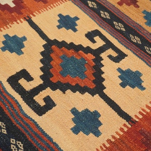 Puede incluir: Primer plano de una alfombra tejida a mano con patrones geom&eacute;tricos. La alfombra presenta un campo central de color beige con dise&ntilde;os negros, azules y &oacute;xido. El borde tiene rayas alternas azules, &oacute;xido y beige.