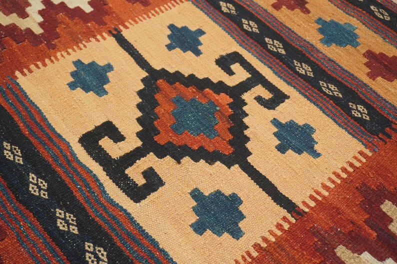 Puede incluir: Primer plano de una alfombra tejida a mano con patrones geom&eacute;tricos. La alfombra presenta un campo central de color beige con dise&ntilde;os negros, azules y &oacute;xido. El borde tiene rayas alternas azules, &oacute;xido y beige.