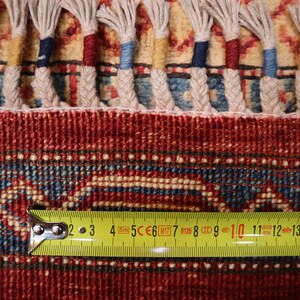 May include: Close-up of a red, blue, and cream colored rug with a fringe border. A yellow measuring tape is placed on the rug, showing measurements from 2 to 15 inches.