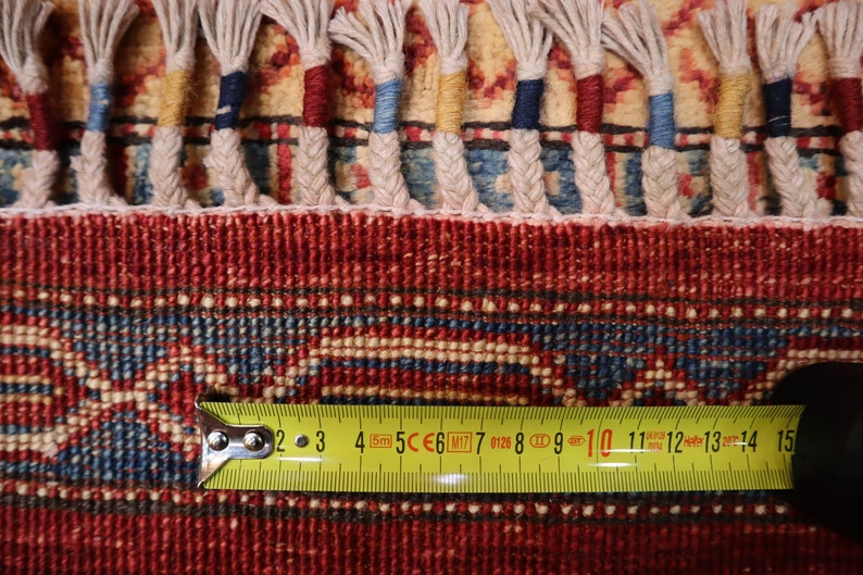 May include: Close-up of a red, blue, and cream colored rug with a fringe border. A yellow measuring tape is placed on the rug, showing measurements from 2 to 15 inches.