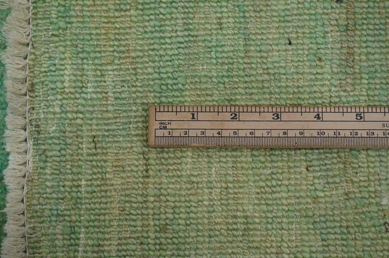 May include: Close-up of a green woven rug with a white fringe. A ruler is placed on the rug to show the texture and weave. The ruler measures 5 inches.