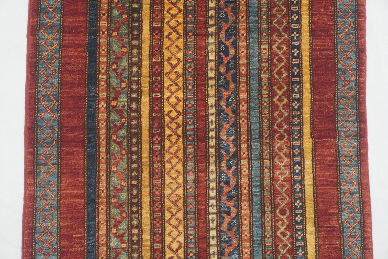 Może przedstawiać: Prostokątny dywan z powtarzającym się wzorem pionowych pask&oacute;w. Dywan ma paski w odcieniach czerwieni, niebieskiego, ż&oacute;łtego i zielonego. Wz&oacute;r zawiera motywy geometryczne i kwiatowe, tworząc teksturowany wygląd. Dywan jest prawdopodobnie wykonany z tkanego materiału.