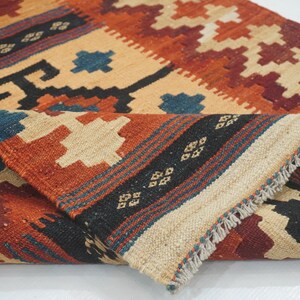 Puede incluir: Una alfombra plegada, tejida a mano, con un patr&oacute;n geom&eacute;trico. La alfombra presenta una paleta de colores beige, naranja, rojo, negro y azul. El dise&ntilde;o incluye diamantes y formas geom&eacute;tricas. La alfombra tiene un borde con flecos.
