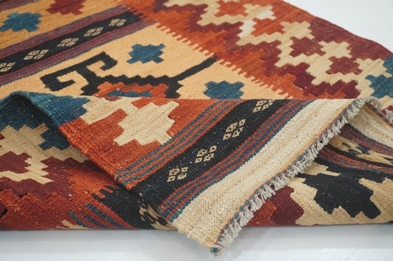 Puede incluir: Una alfombra plegada, tejida a mano, con un patr&oacute;n geom&eacute;trico. La alfombra presenta una paleta de colores beige, naranja, rojo, negro y azul. El dise&ntilde;o incluye diamantes y formas geom&eacute;tricas. La alfombra tiene un borde con flecos.