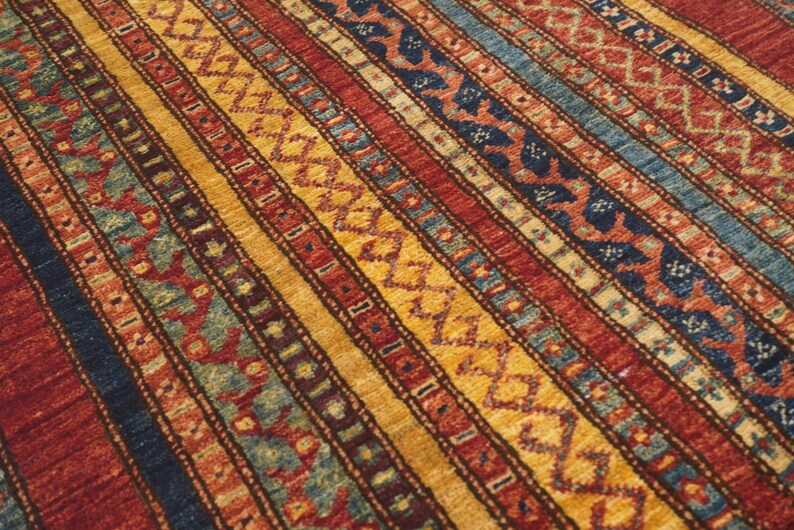 Może przedstawiać: Zbliżenie kolorowego dywanu z wzorem w paski. Dywan ma naprzemienne pasy w kolorze czerwonym, złotym, niebieskim i morskim. Wz&oacute;r zawiera motywy geometryczne i kwiatowe, tworząc teksturowaną i atrakcyjną wizualnie powierzchnię.