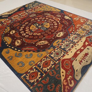 Op de afbeelding: Een kleurrijk tapijt met een geometrisch patroon in tinten blauw, rood, geel en bruin. Het tapijt heeft een centraal medaillon met een cirkelvormig ontwerp en een rand met een herhalend patroon.