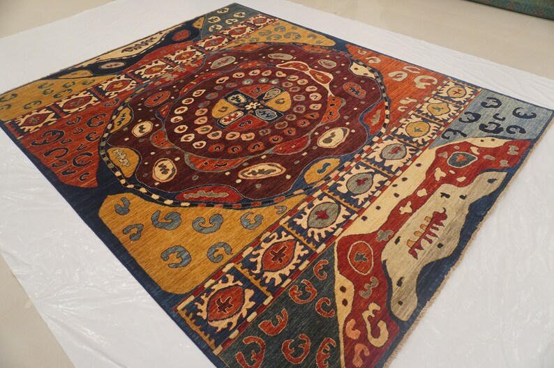Op de afbeelding: Een kleurrijk tapijt met een geometrisch patroon in tinten blauw, rood, geel en bruin. Het tapijt heeft een centraal medaillon met een cirkelvormig ontwerp en een rand met een herhalend patroon.