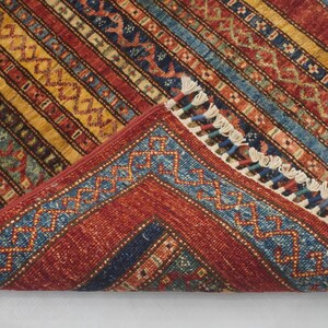 Może przedstawiać: Złożony, prostokątny dywan o szczeg&oacute;łowym wzorze. Dywan ma czerwoną podstawę z ż&oacute;łtymi, niebieskimi i brązowymi paskami. Krawędzie mają niebieską obw&oacute;dkę z geometrycznym wzorem i białymi frędzlami.