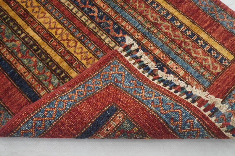 Może przedstawiać: Złożony, prostokątny dywan o szczeg&oacute;łowym wzorze. Dywan ma czerwoną podstawę z ż&oacute;łtymi, niebieskimi i brązowymi paskami. Krawędzie mają niebieską obw&oacute;dkę z geometrycznym wzorem i białymi frędzlami.