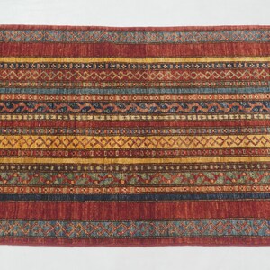 Może przedstawiać: Prostokątny dywan ze wzorem w paski. Dywan ma naprzemienne pasy w kolorze czerwonym, ż&oacute;łtym, niebieskim i brązowym z geometrycznymi wzorami. Dywan jest prawdopodobnie wykonany z tkanej tkaniny i nadaje się do przedpokoju lub wejścia.