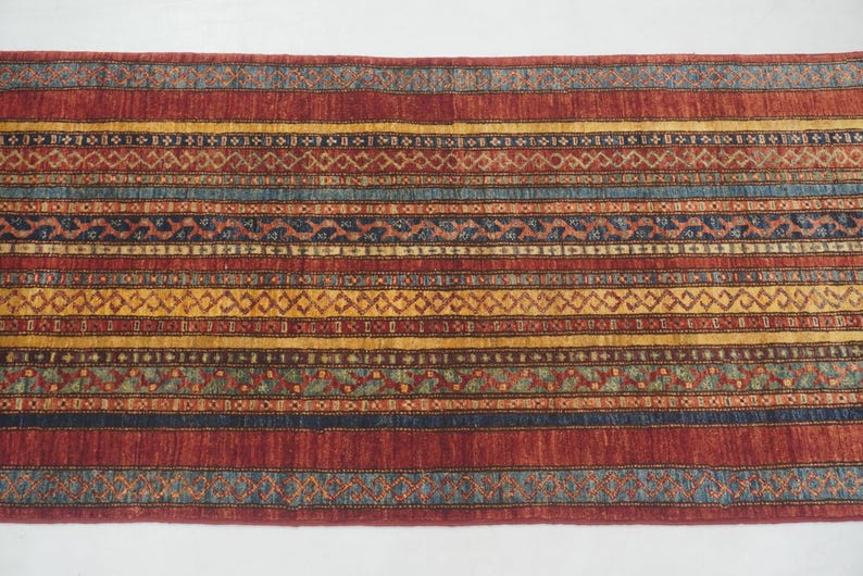 Może przedstawiać: Prostokątny dywan ze wzorem w paski. Dywan ma naprzemienne pasy w kolorze czerwonym, ż&oacute;łtym, niebieskim i brązowym z geometrycznymi wzorami. Dywan jest prawdopodobnie wykonany z tkanej tkaniny i nadaje się do przedpokoju lub wejścia.