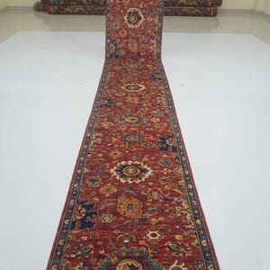 May include: A long, rectangular red runner rug with an intricate floral pattern in shades of blue, orange, and cream. The rug is rolled up at the end, showcasing its length and detailed design. The rug is likely made of wool.