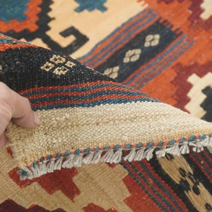 Puede incluir: Una mano levanta una secci&oacute;n de una alfombra tejida, revelando su reverso. La alfombra presenta un patr&oacute;n geom&eacute;trico en tonos beige, rojo, negro y turquesa. Los bordes tienen flecos. El dise&ntilde;o de la alfombra es visible.