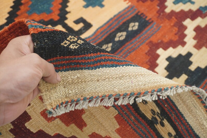 Puede incluir: Una mano levanta una secci&oacute;n de una alfombra tejida, revelando su reverso. La alfombra presenta un patr&oacute;n geom&eacute;trico en tonos beige, rojo, negro y turquesa. Los bordes tienen flecos. El dise&ntilde;o de la alfombra es visible.