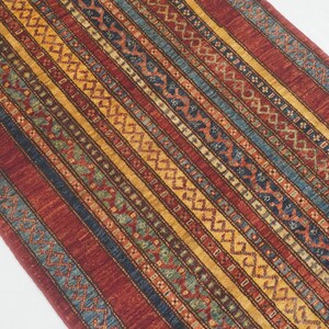 Może przedstawiać: Prostokątny dywan ze wzorem w paski w odcieniach czerwieni, ż&oacute;łci, niebieskiego i zieleni. Dywan posiada geometryczne wzory i teksturowaną powierzchnię, odpowiedni do dekoracji wnętrz.