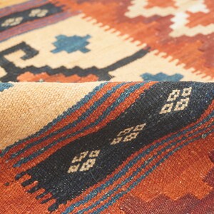 Puede incluir: Primer plano de una alfombra tejida con un borde doblado. La alfombra presenta un patr&oacute;n geom&eacute;trico en tonos beige, &oacute;xido, negro y azul. El dise&ntilde;o incluye formas de diamantes y zigzags, creando una apariencia texturizada.