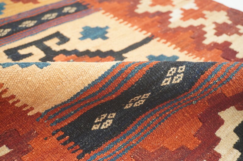 Puede incluir: Primer plano de una alfombra tejida con un borde doblado. La alfombra presenta un patr&oacute;n geom&eacute;trico en tonos beige, &oacute;xido, negro y azul. El dise&ntilde;o incluye formas de diamantes y zigzags, creando una apariencia texturizada.