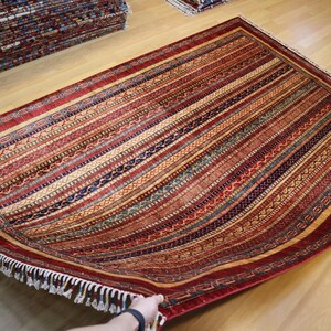 May include: A red, blue, and gold patterned rug with a fringe border. The rug is folded in half and being held by a hand.