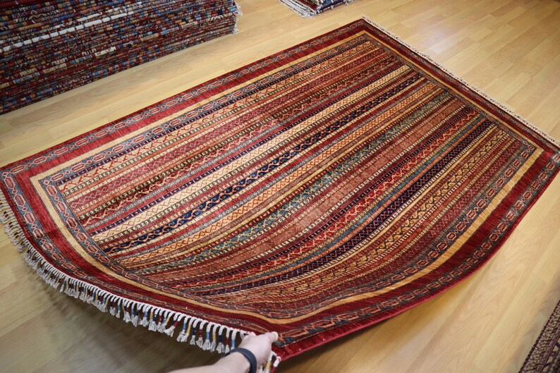 May include: A red, blue, and gold patterned rug with a fringe border. The rug is folded in half and being held by a hand.