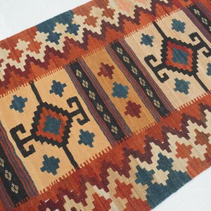 Puede incluir: Una alfombra rectangular tejida con un patr&oacute;n geom&eacute;trico. La alfombra presenta un campo central de color beige con formas geom&eacute;tricas repetidas en azul oscuro, rojo y negro. Los bordes son una mezcla de zigzags rojos, naranjas y crema. La alfombra es probablemente de lana.