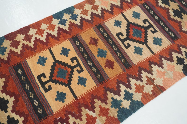 Puede incluir: Una alfombra rectangular tejida con un patr&oacute;n geom&eacute;trico. La alfombra presenta un campo central de color beige con formas geom&eacute;tricas repetidas en azul oscuro, rojo y negro. Los bordes son una mezcla de zigzags rojos, naranjas y crema. La alfombra es probablemente de lana.