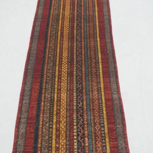 Może przedstawiać: Długi, prostokątny dywan z czerwonym tłem i pionowymi paskami w kolorze ż&oacute;łtym, niebieskim i brązowym. Dywan ma wzorzystą niebieską obw&oacute;dkę i frędzle na końcach. Dywan jest prawdopodobnie wykonany z tkanego materiału.