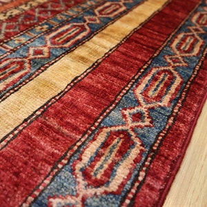 May include: Close-up of a red, blue, and beige patterned rug with intricate designs. The rug has a fringe border and is made of a soft, woven material.
