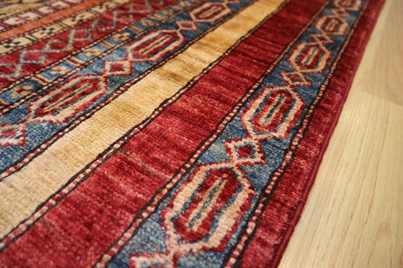 May include: Close-up of a red, blue, and beige patterned rug with intricate designs. The rug has a fringe border and is made of a soft, woven material.