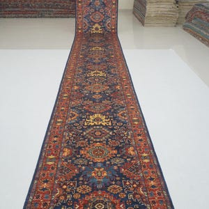 May include: A long, narrow runner rug with an intricate, geometric pattern. The rug features a deep blue background with red, orange, and yellow accents. The design is symmetrical, with a central medallion and repeating motifs along the borders. The rug is likely made of woven material.