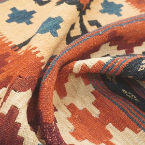 Puede incluir: Primer plano de una alfombra plegada con un dise&ntilde;o geom&eacute;trico. La alfombra presenta un fondo color crema con acentos en tonos naranja, burdeos, azul y negro. El dise&ntilde;o incluye patrones de diamantes y zig-zag.