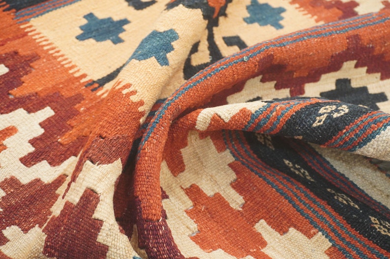 Puede incluir: Primer plano de una alfombra plegada con un dise&ntilde;o geom&eacute;trico. La alfombra presenta un fondo color crema con acentos en tonos naranja, burdeos, azul y negro. El dise&ntilde;o incluye patrones de diamantes y zig-zag.