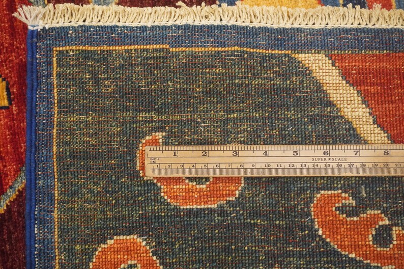 Op de afbeelding: Close-up van een blauw tapijt met een cr&egrave;mekleurige rand en een liniaal die 20 cm meet. Het tapijt heeft een herhalend patroon van oranje en cr&egrave;mekleurige vormen.