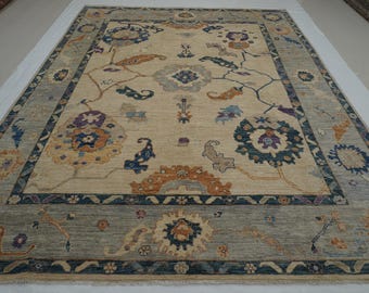 May include: A large, rectangular area rug with a central beige field featuring floral and vine motifs in shades of blue, orange, and purple. The rug has a blue border with similar floral patterns. The overall design is intricate and detailed.