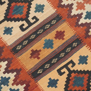Puede incluir: Alfombra tejida a mano con un patr&oacute;n geom&eacute;trico en tonos beige, naranja, marr&oacute;n y azul. La alfombra presenta motivos repetidos de diamantes y cuadrados, creando una superficie texturizada. El dise&ntilde;o incluye rayas y formas estilizadas.