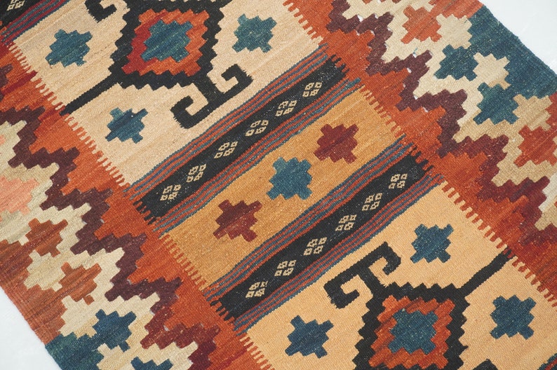 Puede incluir: Alfombra tejida a mano con un patr&oacute;n geom&eacute;trico en tonos beige, naranja, marr&oacute;n y azul. La alfombra presenta motivos repetidos de diamantes y cuadrados, creando una superficie texturizada. El dise&ntilde;o incluye rayas y formas estilizadas.