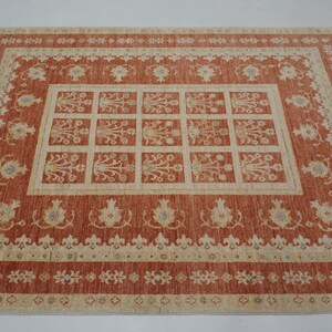 May include: A traditional rug with a red background and a beige pattern. The rug has a central design of a grid of squares with floral motifs. The border of the rug is decorated with a repeating pattern of small flowers.